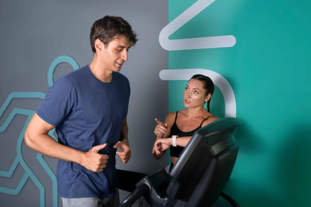 A fitness trainer guiding a man jogging on a treadmill in a modern gym setting.