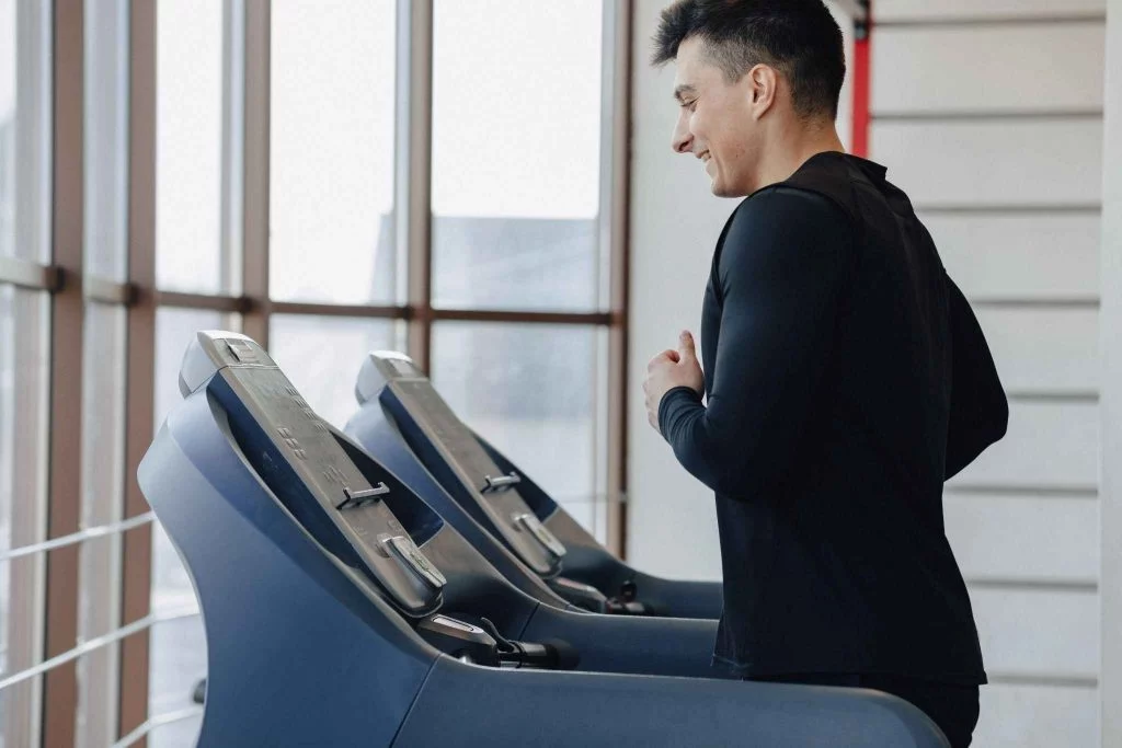 A man performing a treadmill workout beside large gym windows.
