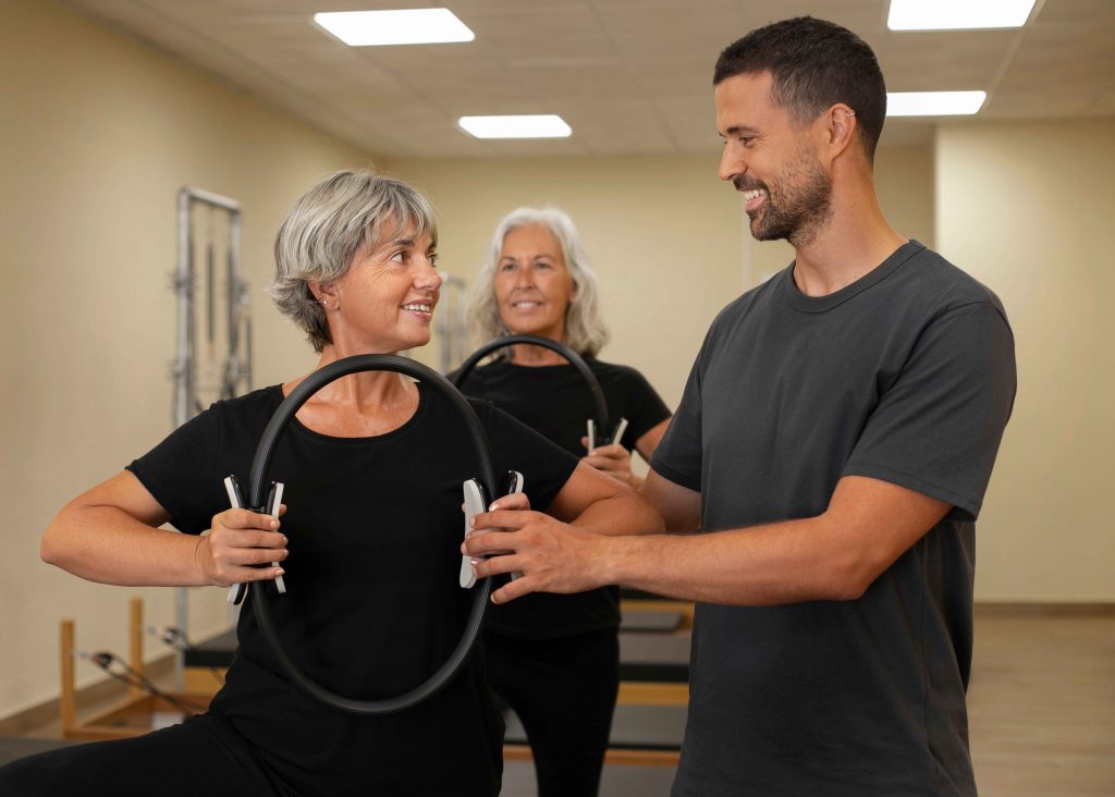 front-view-people-pilates-class_11zon Senior women working with a male personal trainer for seniors, using a Pilates ring to improve balance and strength.