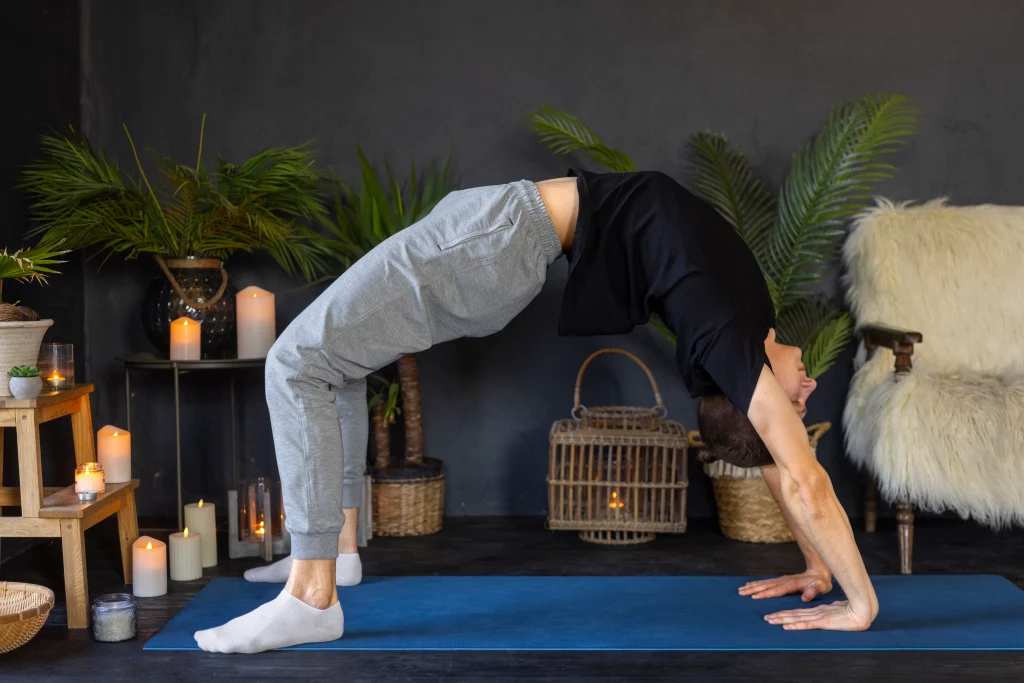 Man performing a backbend yoga pose on a mat indoors with candles and plants in the background.