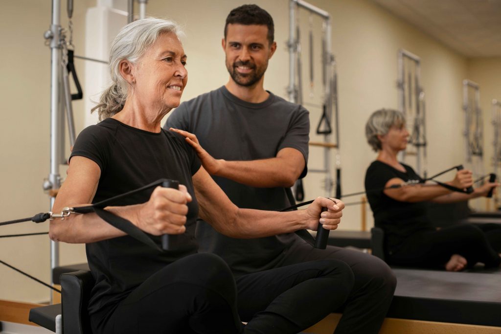 side-view-people-pilates-class_11zon Senior women working with male personal trainers for seniors, using a Pilates ring to improve balance and strength.
