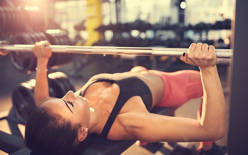 A young girl lying on a bench with a barbell trying a basic bench press as a compound chest exercise