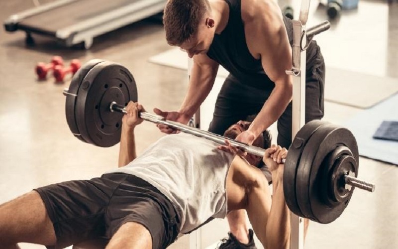 A man lying on a bench trying to do chest exercise with the aid of his coach