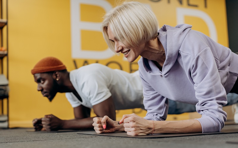 A blonde middle-aged women and a black man are in a plank position 