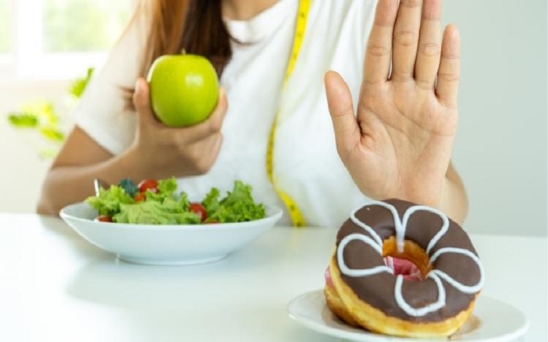 A woman holding a green apple in her hand saying no to a chocolate doughnut 