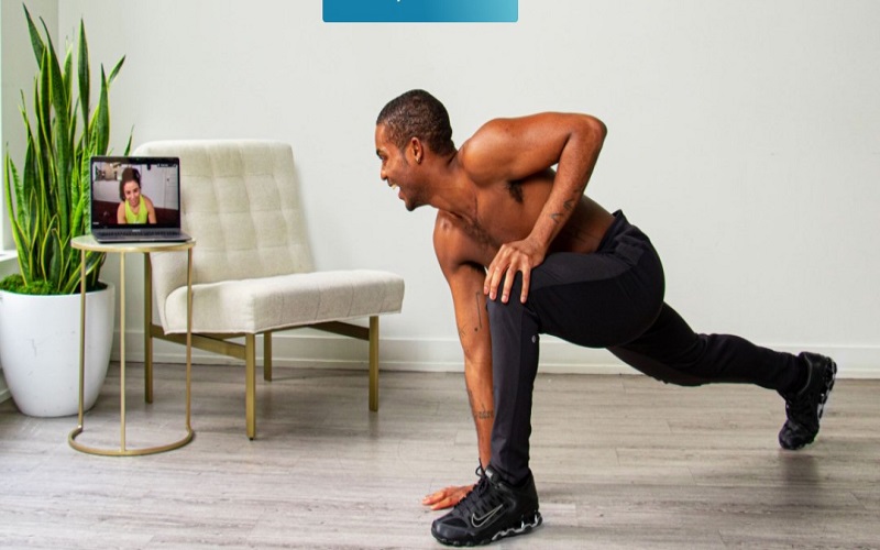 A man is stretching his leg, and working out while watching his online personal trainer on a laptop 