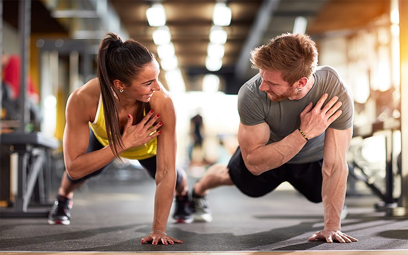 A man and a woman are doing one-handed pushup at a gym. 