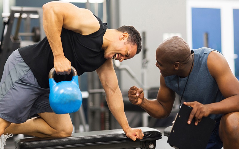 A black personal trainer is training a man trying a heavy kettle ball