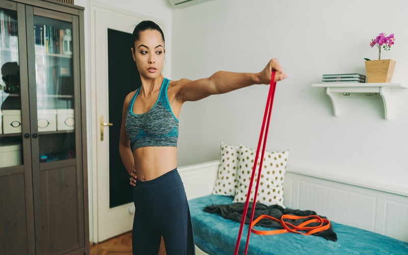 A woman is working on her chest isolation exercise with a simple resistance band at home. She is pulling the band with her left hand, while at a shoulder-length wide position and the right hand is on the wrist 