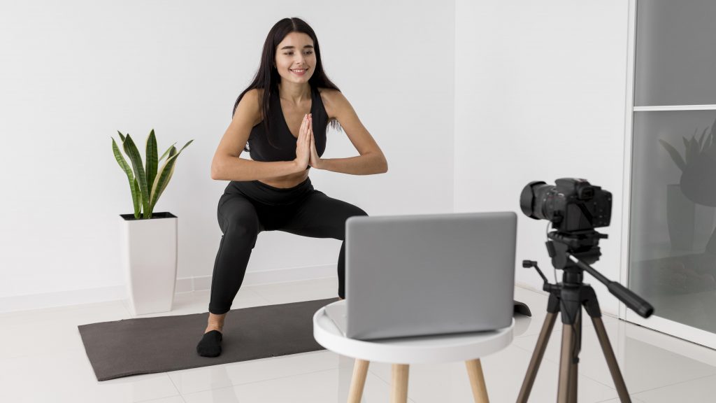 female-influencer-home-vlogging-while-exercising A virtual female personal trainer demonstrates a squat while coaching a client through an online workout session via laptop.