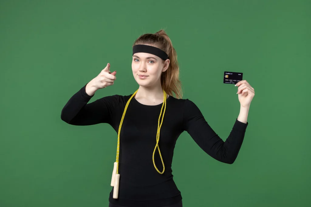Young woman in fitness clothing holding a credit card while posing confidently against a green background.