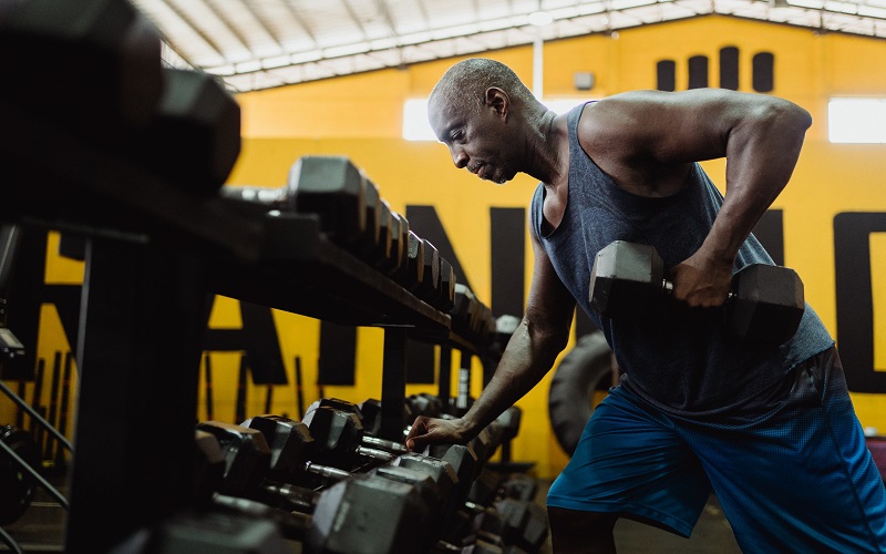 A black man is at the gym working out on his massive biceps and triceps with dumbbells