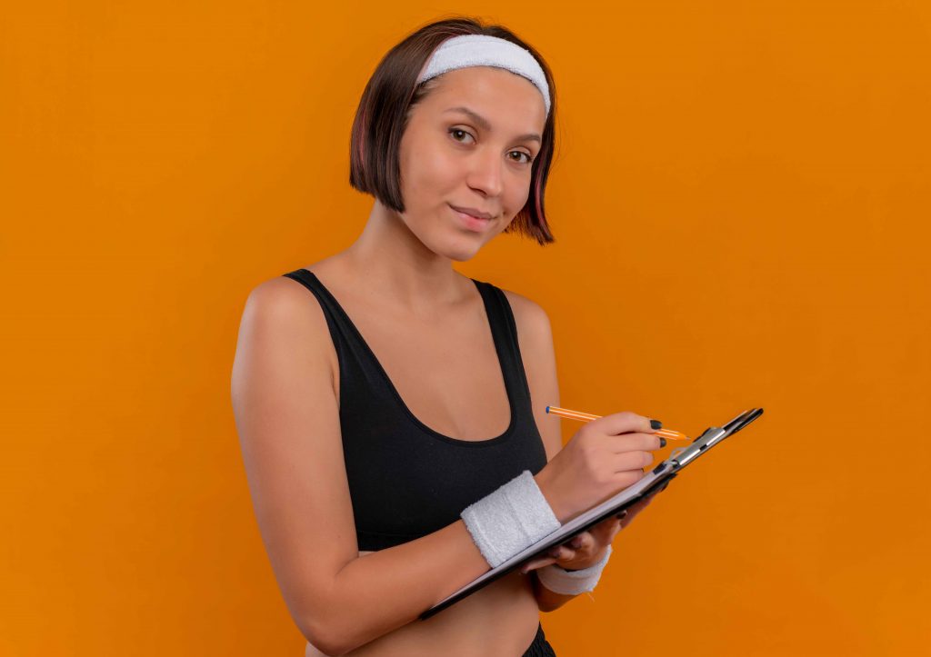 young-fitness-woman-sportswear-with-headband-holding-clipboard-with-blank-pages-writing-looking-confident-standing-orange-wall_11zon Personal trainers cost highlighted by a professional trainer holding a clipboard confidently.