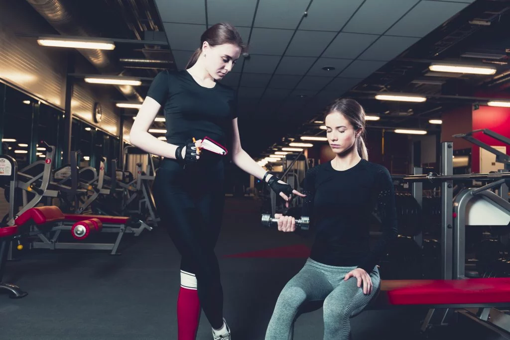 female-trainer-assisting-woman-while-exercising-with-dumbbell A female personal trainer assisting a client with dumbbell exercises, showcasing personalized fitness training services.