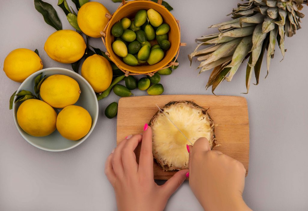 top-view-female-hands-cutting-pineapple-wooden-kitchen-board-with-knife-with-lemons-bowl-with-kinkans-bucket-white-surface_11zon Hands cutting pineapple, a fresh start for Pineapple for Weight Loss meals.