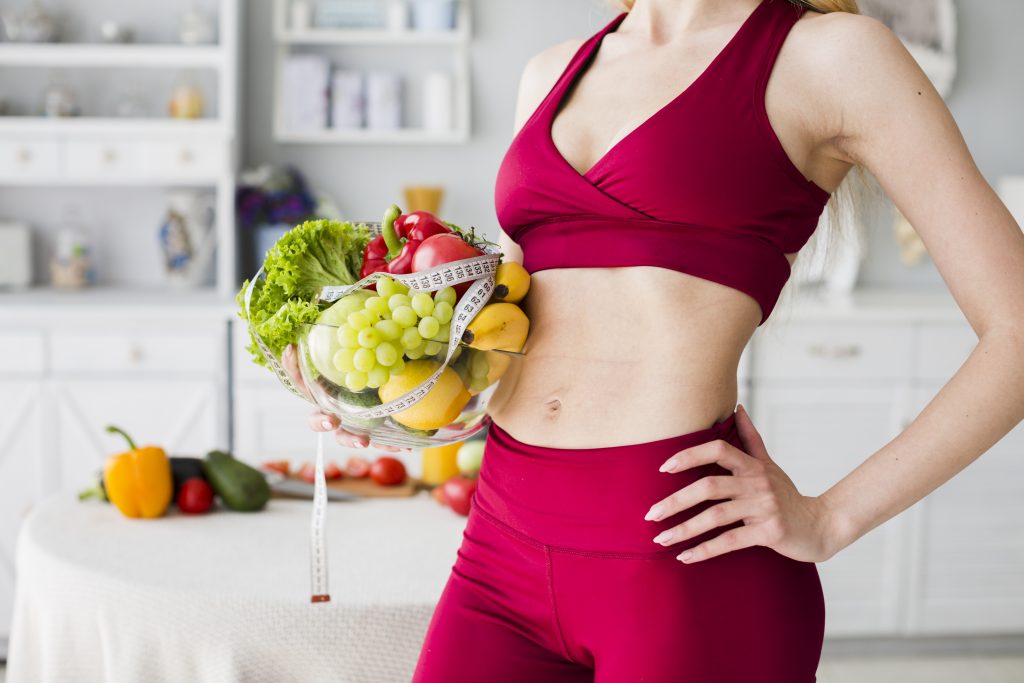 Sporty woman in fitness wear holding a bowl of fresh fruits and vegetables wrapped with a measuring tape, symbolizing healthy eating for weight loss.