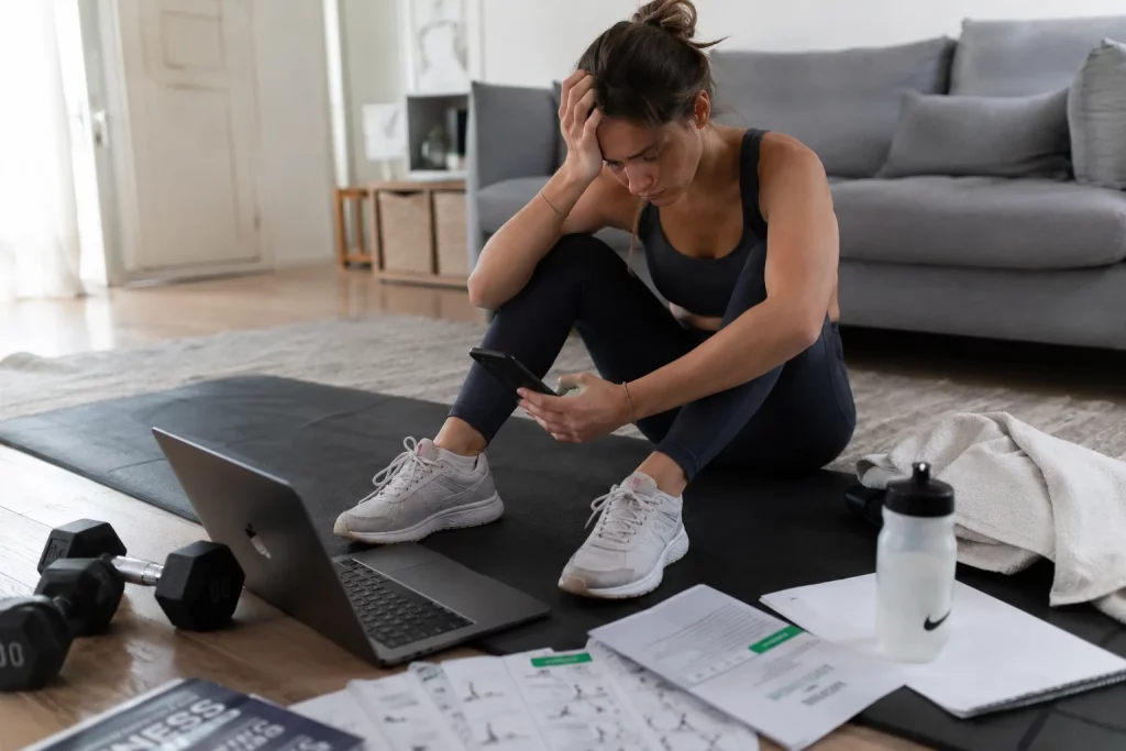 Most Expensive “Cheap” Mistakes to Avoid A woman pausing during her workout to compare online personal trainer rates on her phone.
