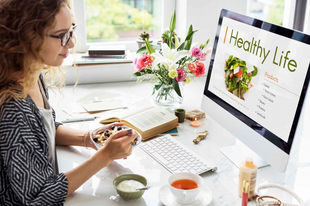A woman is enjoying a healthy meal while browsing a nutrition website on her computer, with flowers and books on the desk.