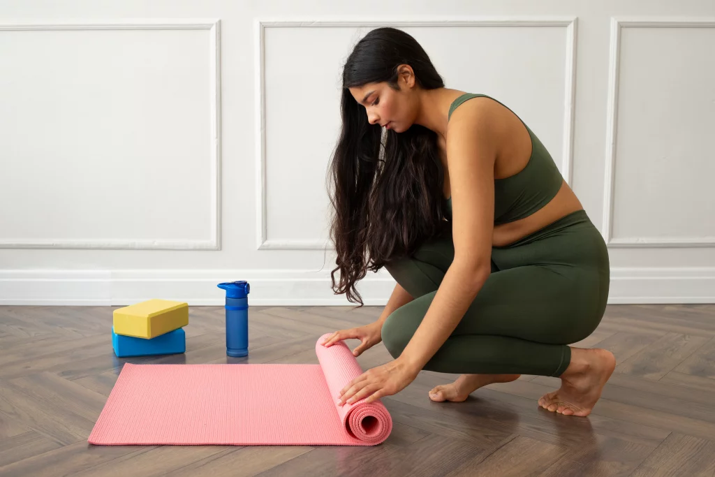 Yoga for beginners preparation at home with a woman rolling out a pink yoga mat beside yoga blocks and a blue water bottle on a wooden floor.