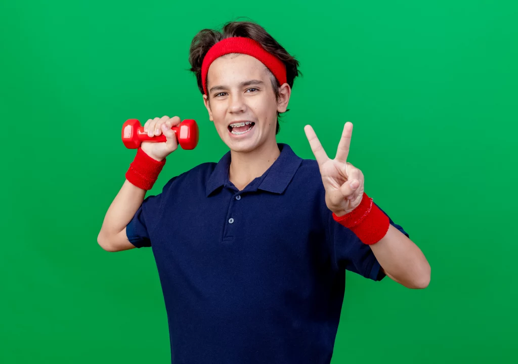 fitness trainer for teens. A cheerful teenage boy holding a red dumbbell and posing after an online workout with his fitness trainer for teens.