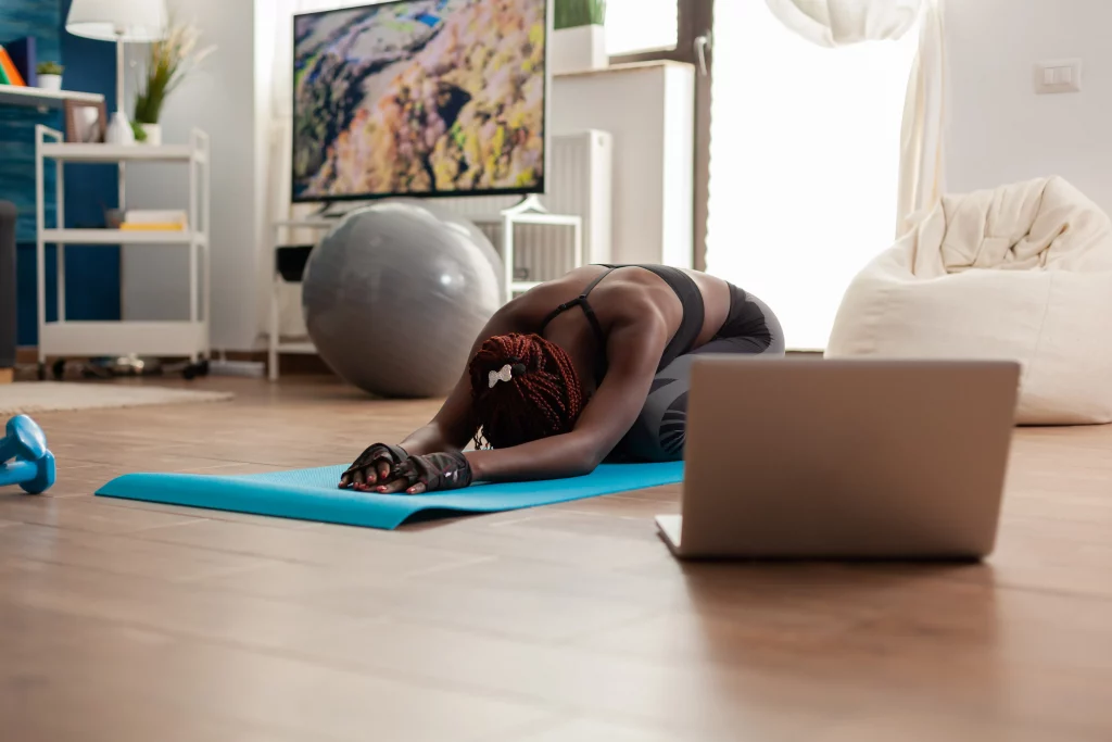 Online General Fitness Trainers A woman stretching on a yoga mat at home while following an online session with general fitness trainers on a laptop.