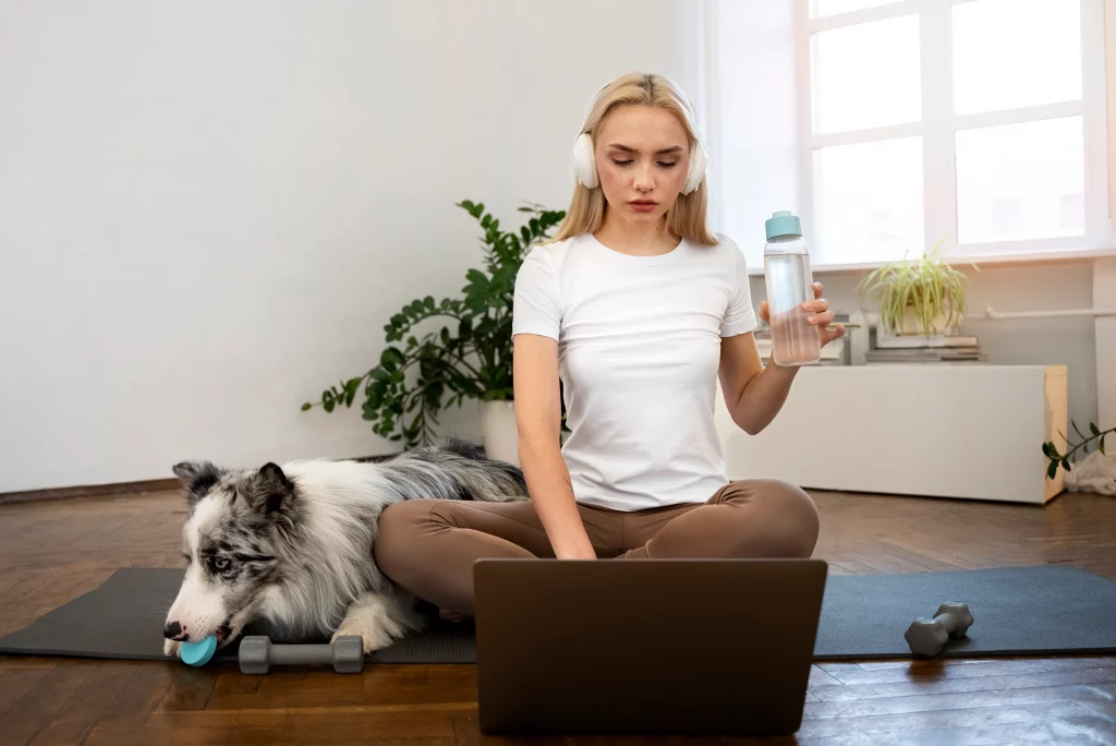 A woman exercising at home while following guidance from affordable online personal trainers on her laptop, with her pet by her side.