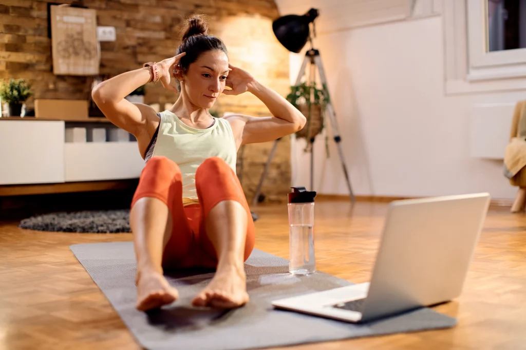 Fit woman doing sit-ups at home while following a Virtual personal training session on her laptop