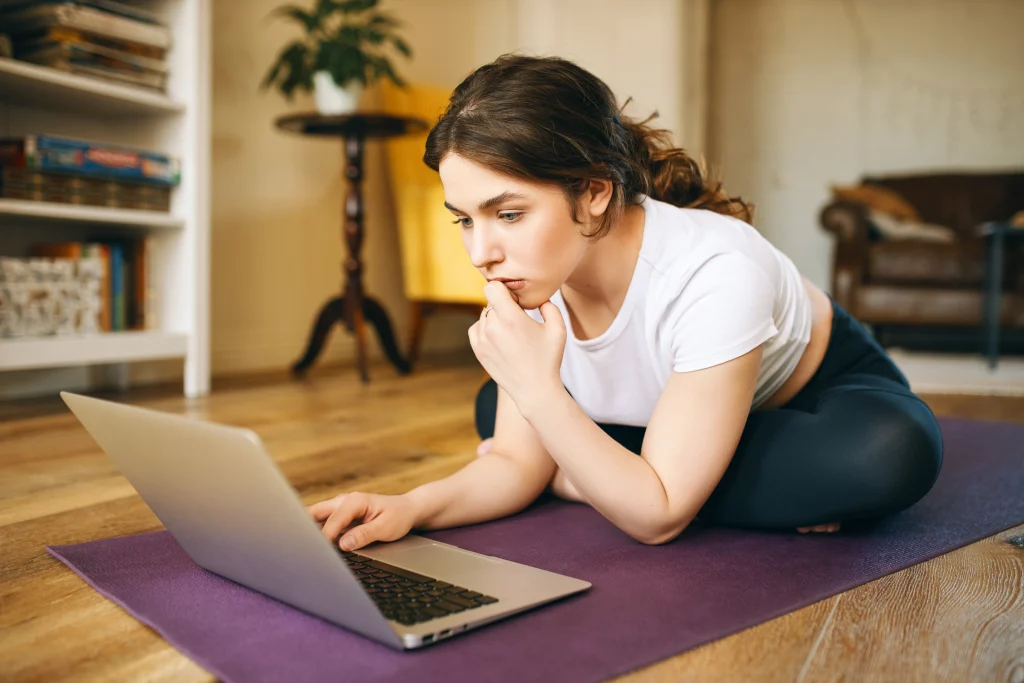 attractive-young-professinal-female-fitness-trainer-sitting-mat-with-portable-computer-having-pensive-look-recording-video-class-online-training-pretty-girl-watching-yoga-tutorial-laptop Woman comparing online yoga classes at home while looking for private yoga instructors through her laptop.