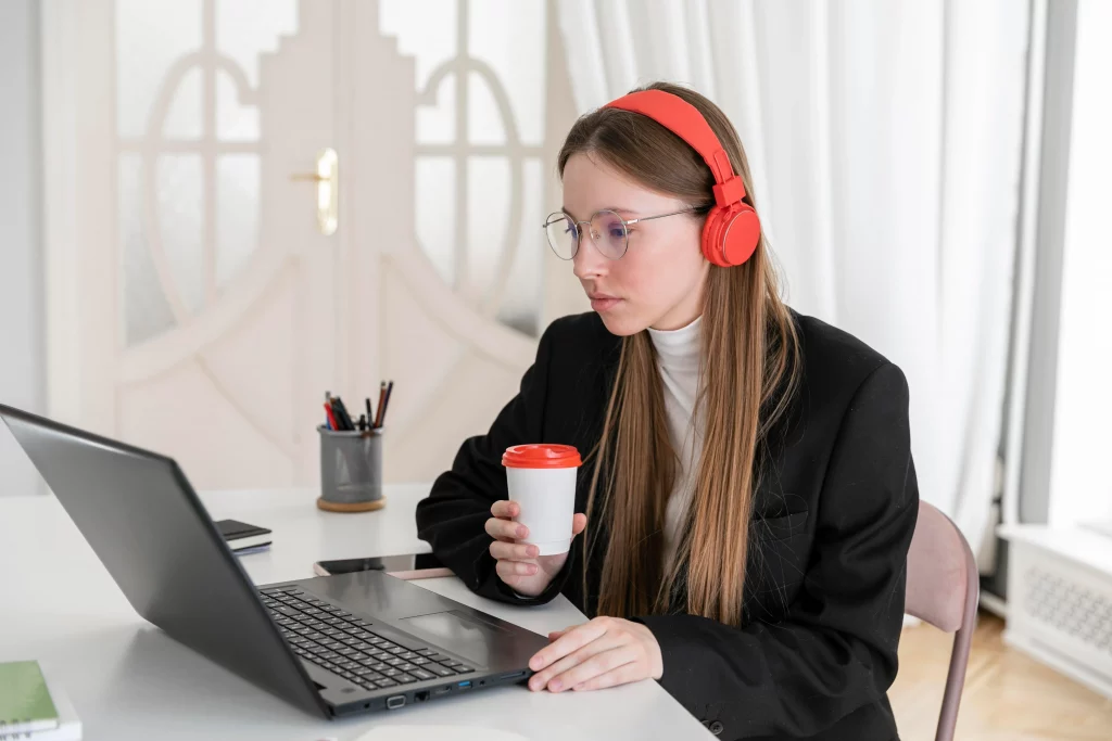 A woman with headphones is searching for online weight loss coaches on her laptop while drinking coffee.