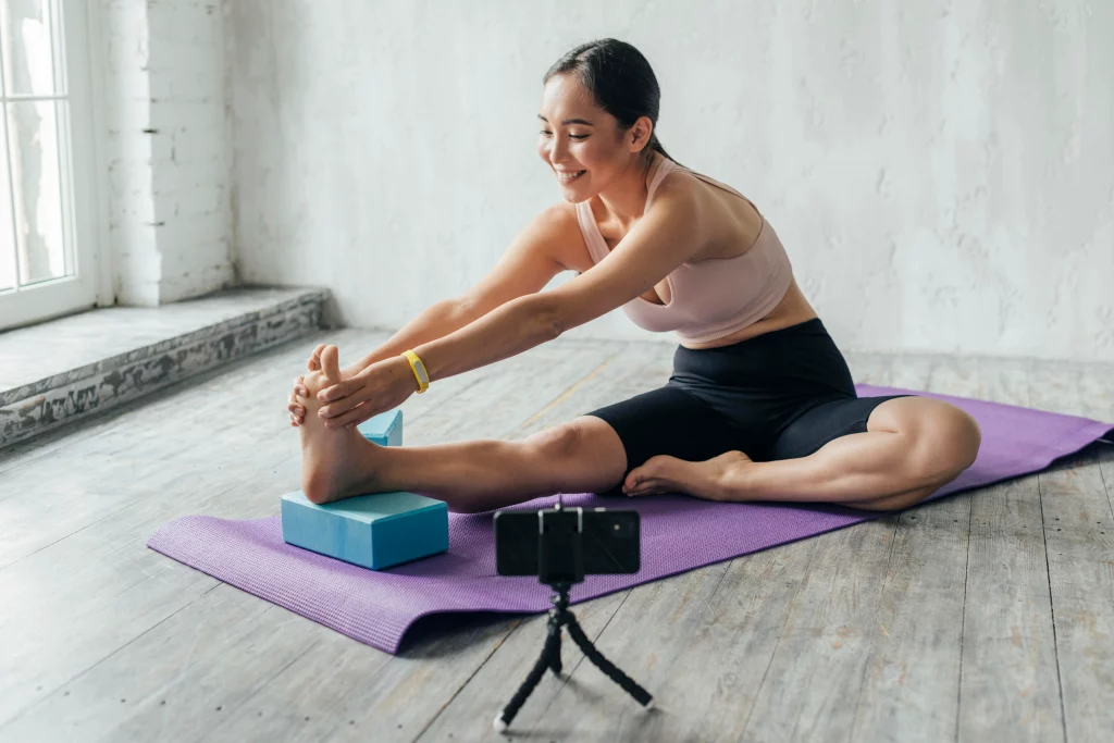 young-woman-vlogging-sportswear-home Woman practicing a flexibility-focused online yoga session at home while following a virtual yoga instructor through her phone.