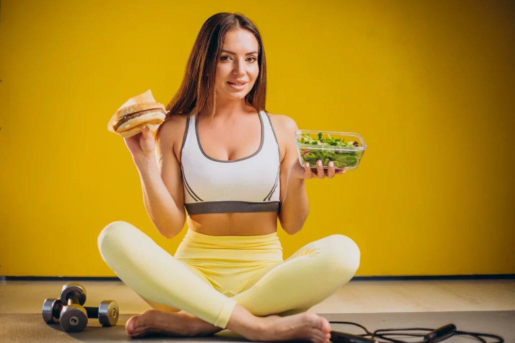 A woman sitting on a workout mat with dumbbells, choosing between a salad and a burger for muscle growth nutrition.