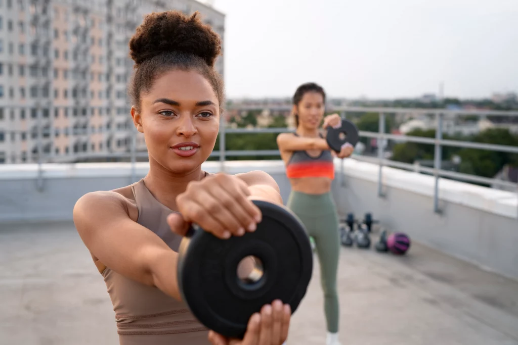 Two women doing strength training exercises with weight plates at a rooftop gym as part of a muscle-building program. 