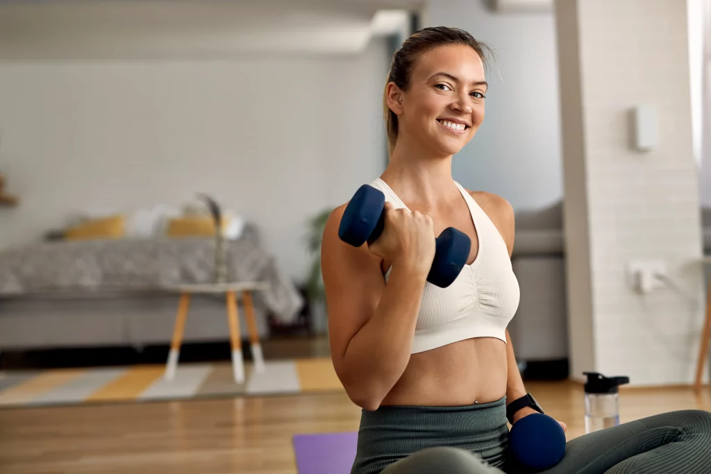 young-happy-sportswoman-exercising-with-dumbbells-home-looking-camera A woman training with dumbbells at home based on a workout plan from a women's fitness app