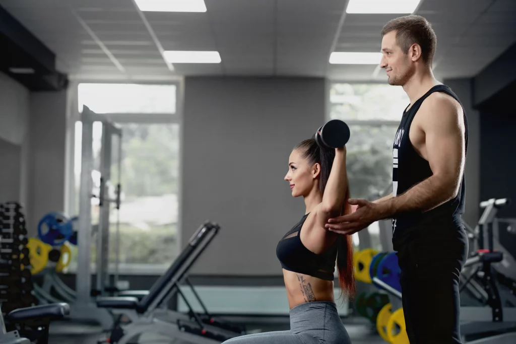 male-personal-trainer-helping-woman-working-with-heavy-dumbbells-gym Personal trainer guiding a woman through a dumbbell workout at one of the top gyms in New York City