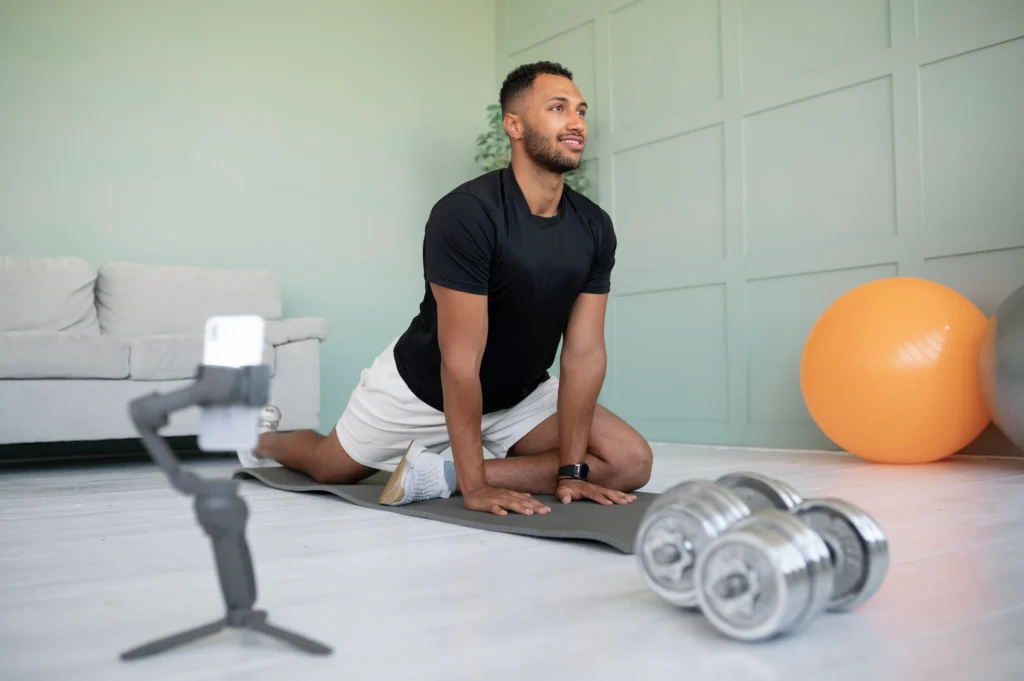 Man stretching on an exercise mat at home with dumbbells and a smartphone, following guidance from a personal trainer for gaining weight.